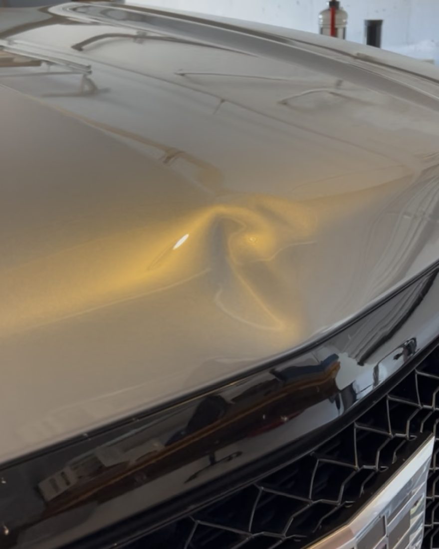 Before: a large dent in the hood of a silver Cadillac Escalade twists the reflection