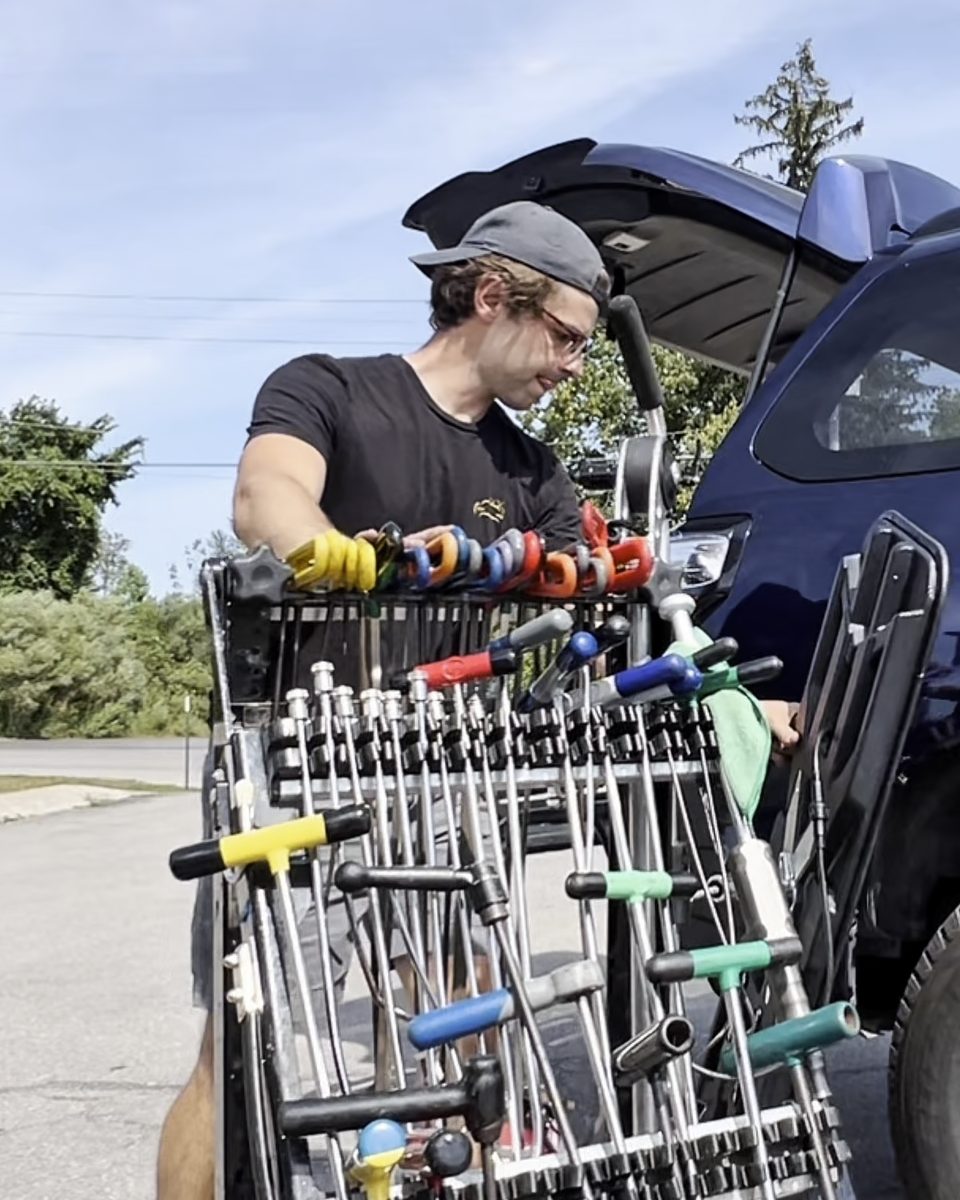 Ethan with his cart of colored dent rods beside an SUV in a sunny driveway